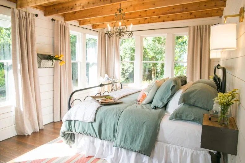Warm exposed wood bedroom ceiling above pale wall boards, soft green bedding and long neutral curtains