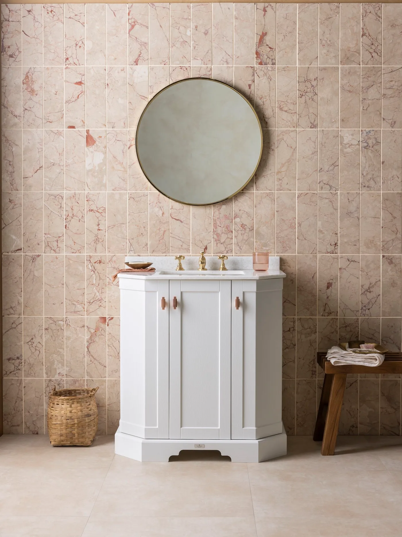 White compact vanity with round mirror against pink veined tile wall beside basket and wood stool