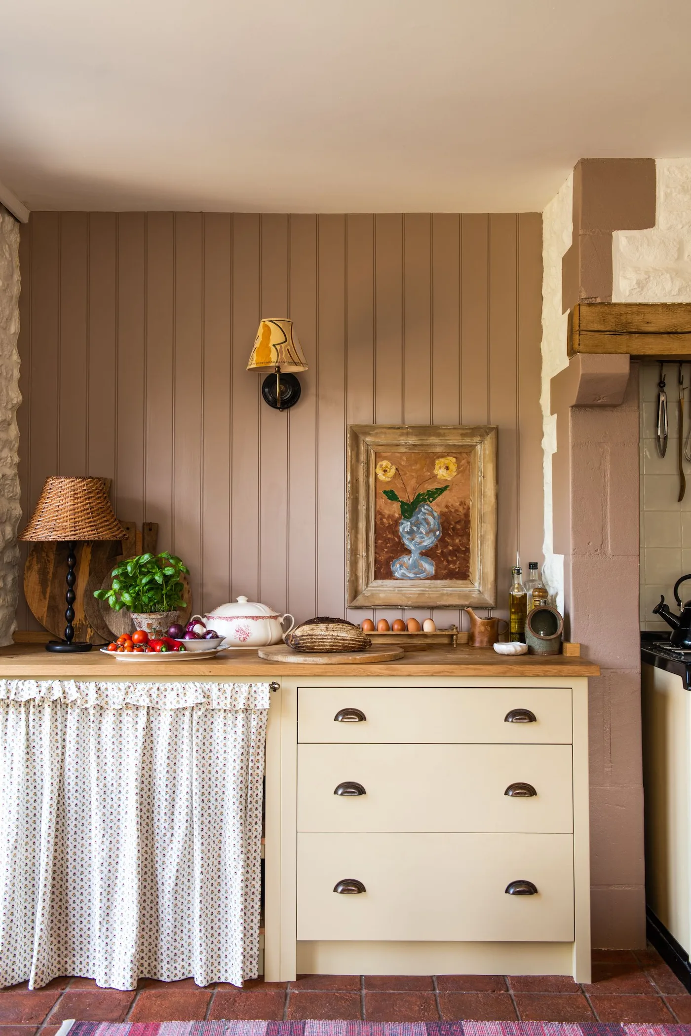 Pale kitchen drawers beside a dotted under-counter curtain, paneled wall, framed flowers and a table lamp.