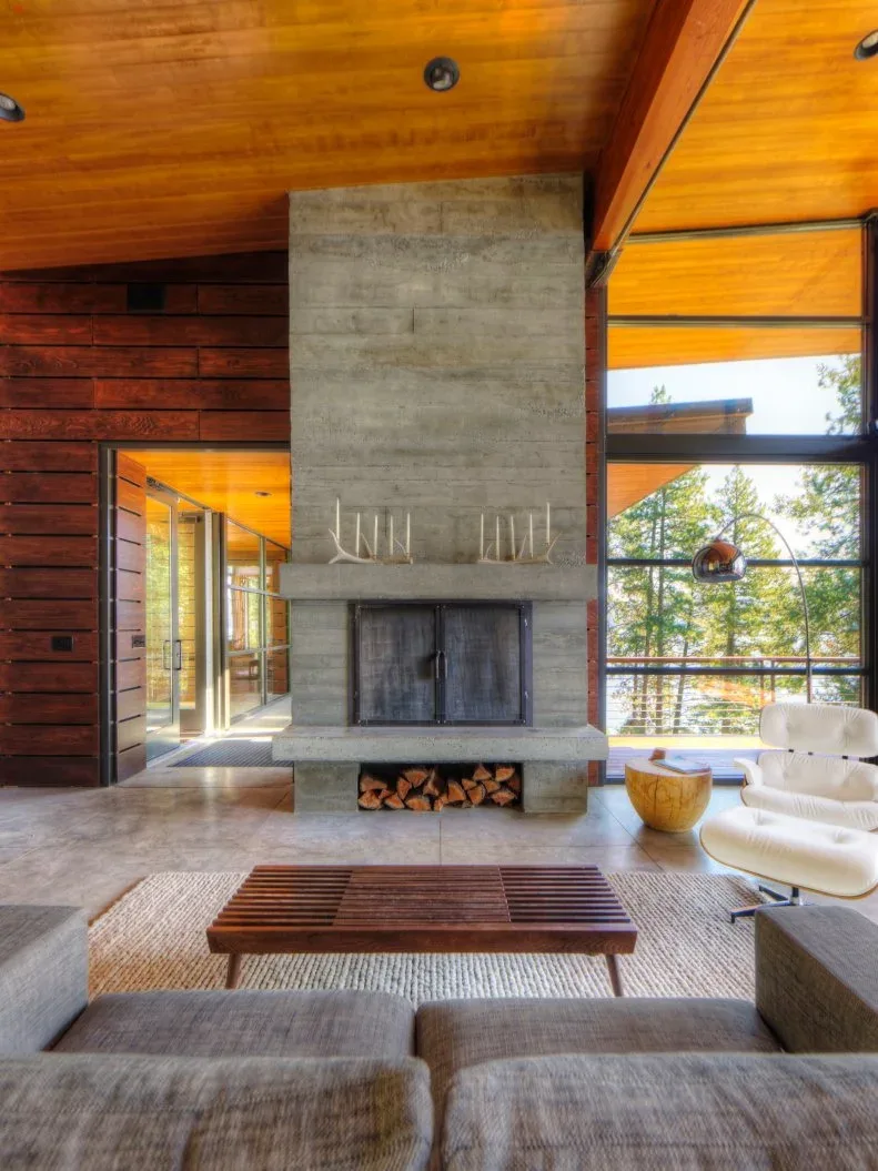 Tall gray block-faced fireplace with low log recess beside warm wood walls, ceiling and large windows