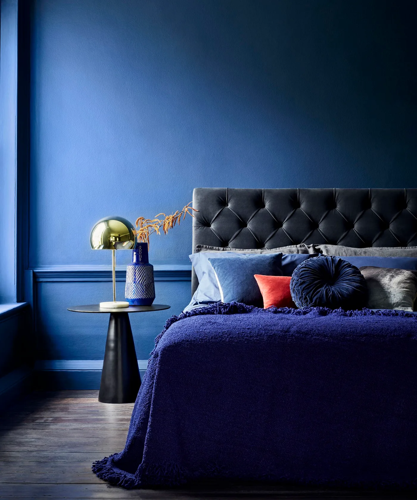 Deep blue bedroom wall and bedding with a dark tufted headboard, an orange cushion, and a domed bedside lamp.