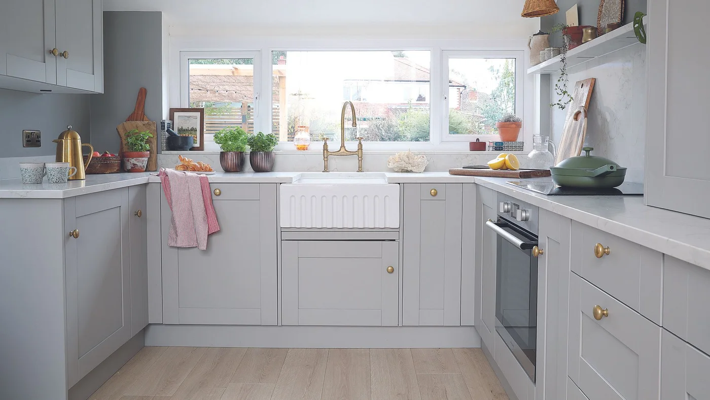 Pale gray cabinets and gold-toned knobs surround a white apron-front sink below a broad window beside a built-in oven