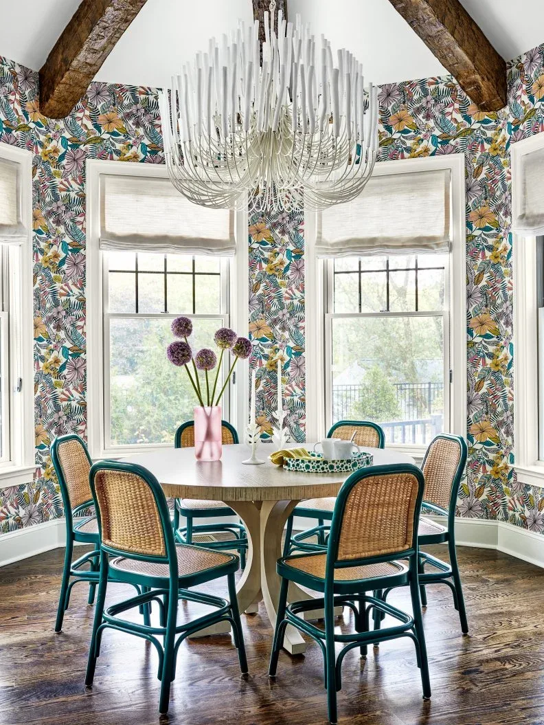 Pale folded shades on black-framed dining windows surrounded by colorful leafy wallpaper and exposed beams