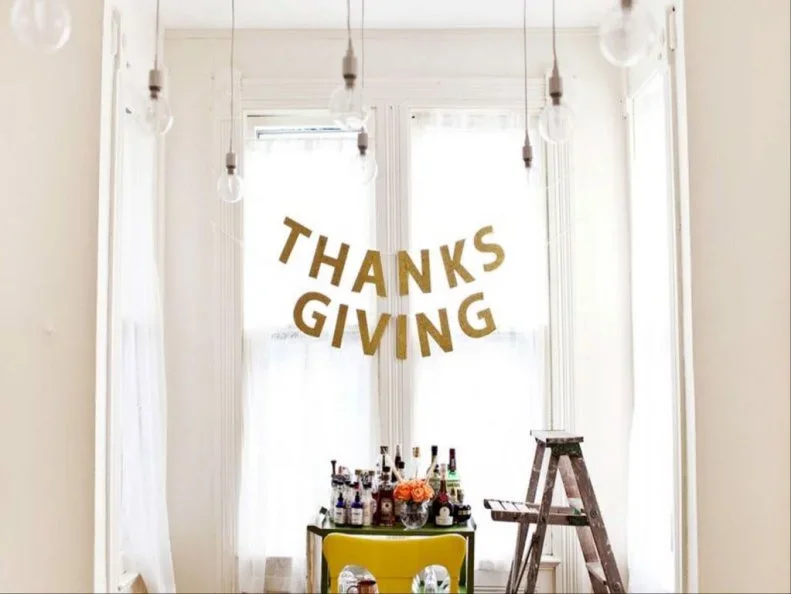 Warm metallic-looking letters spelling Thanks Giving hang in two lines above a small drinks area beside a tall window