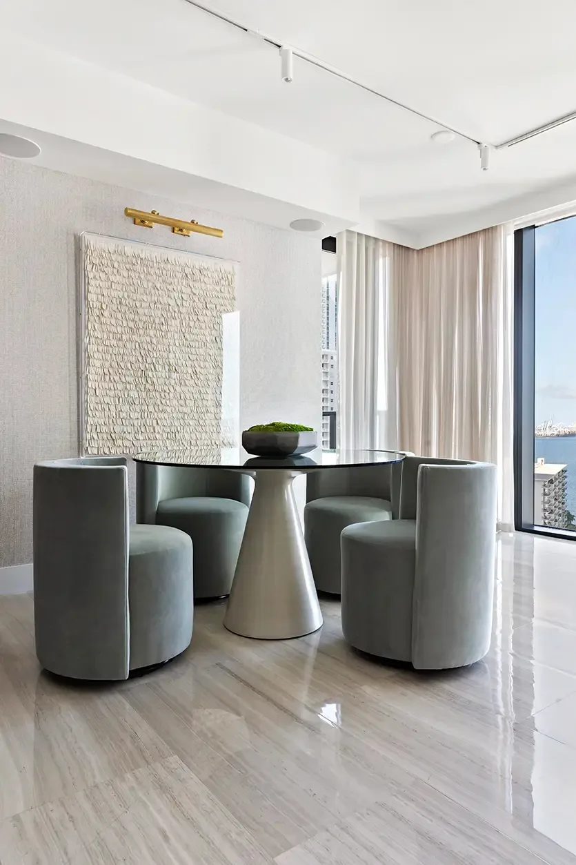 Round dark glass dining table on a pale pedestal surrounded by gray curved chairs beside a window