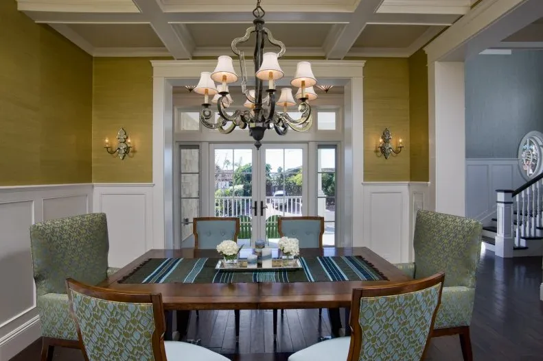 Gold-toned walls above white paneling around a wooden dining table beneath a coffered ceiling and shaded chandelier.