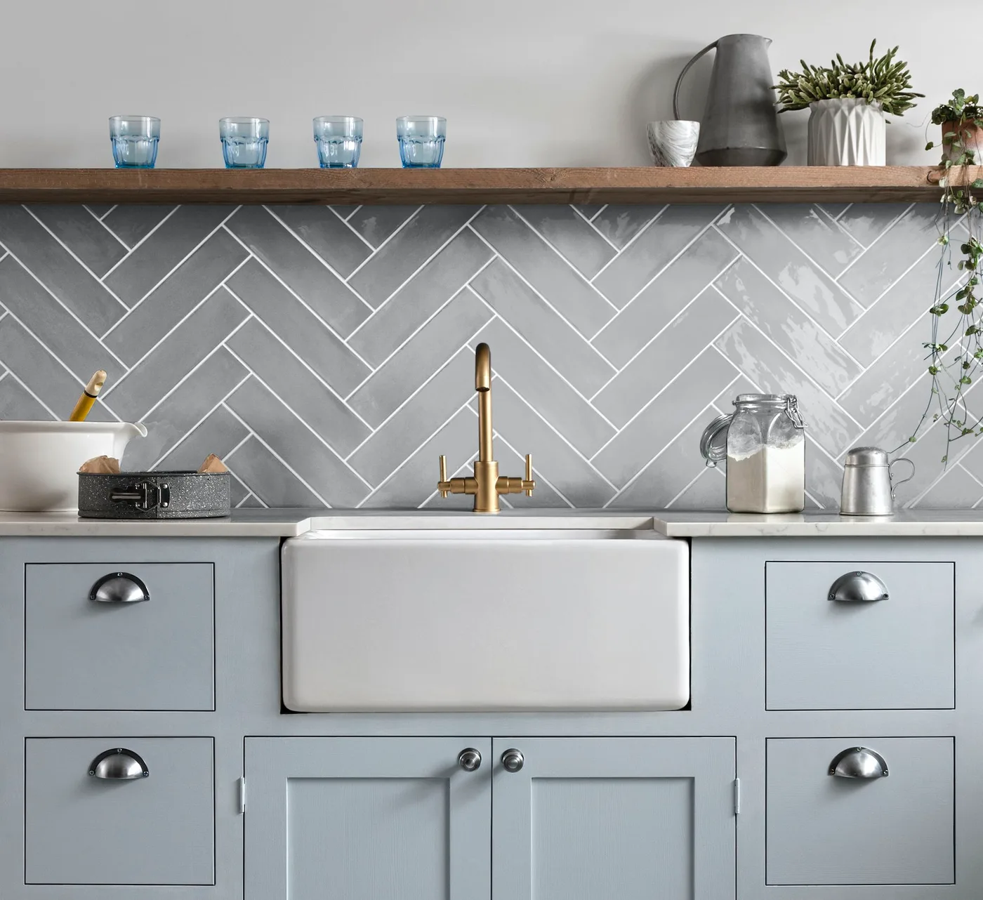 Cottage sink detail with gray framed cabinets, white apron-front sink, warm colored tap, gray herringbone backsplash and long wooden shelf.