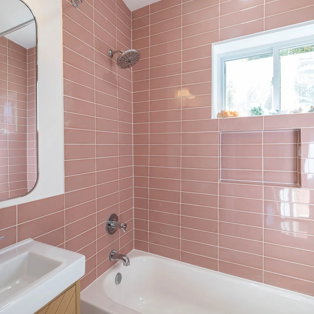 Pink rectangular tiles form a stacked grid around a white bath beneath a small window and beside a recessed niche