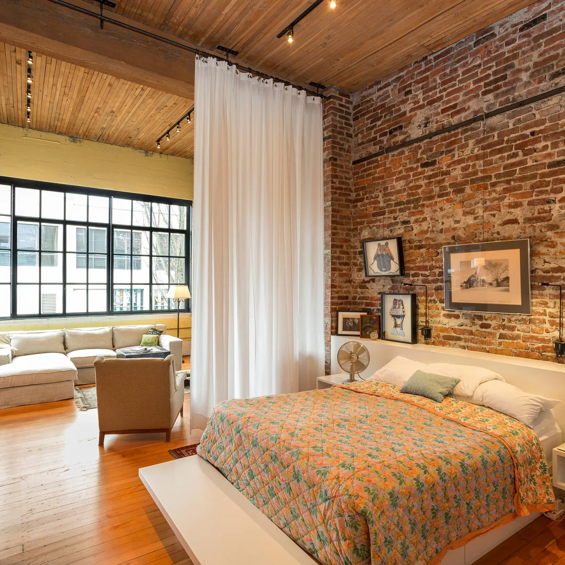 A bed with a colorful floral cover sits against exposed brick beside a white curtain dividing it from a living area with a grid window