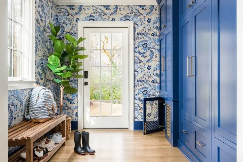 Mudroom with blue cupboards, blue floral wallpaper, wooden bench, woven plant basket and glazed white door.