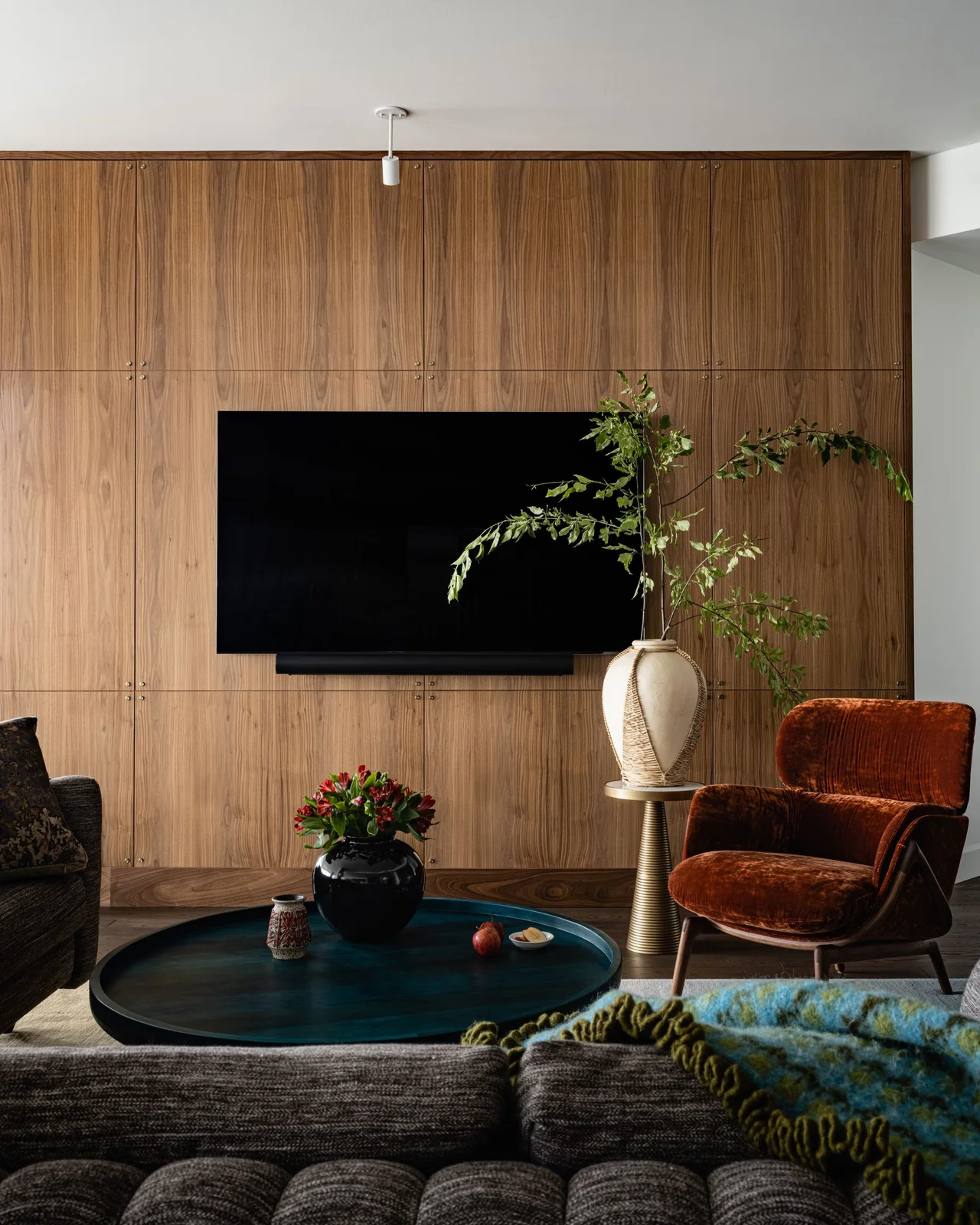 A television and soundbar hang against a full wooden cabinet wall beside a red-brown chair and low round table