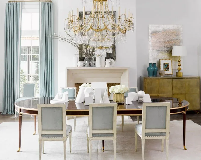 White sculptural objects and low pale flowers on a dark oval dining table with pale upholstered chairs