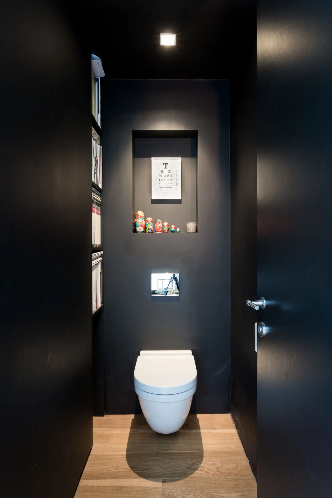 A white toilet stands beneath a small wall recess with artwork and small figures in a narrow dark room beside inset book shelving and a wood floor