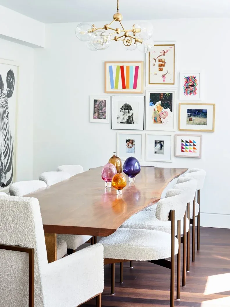 Dining room with a varied colorful gallery on a white wall, a long wooden table, pale textured chairs, colored glass vessels, and a globe chandelier.