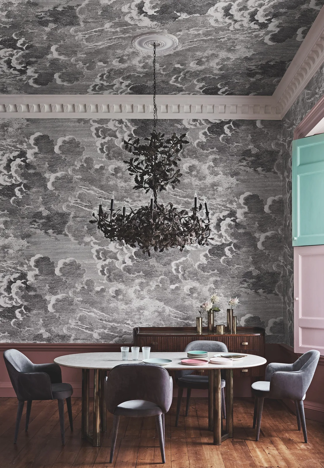 Monochrome cloud-pattern wallpaper covers dining room walls and ceiling above a pale oval table and gray chairs beneath an ornate black chandelier