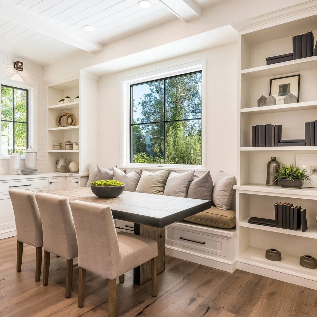 White open shelves and lower cabinets around a dining window bench with a dark table and pale chairs
