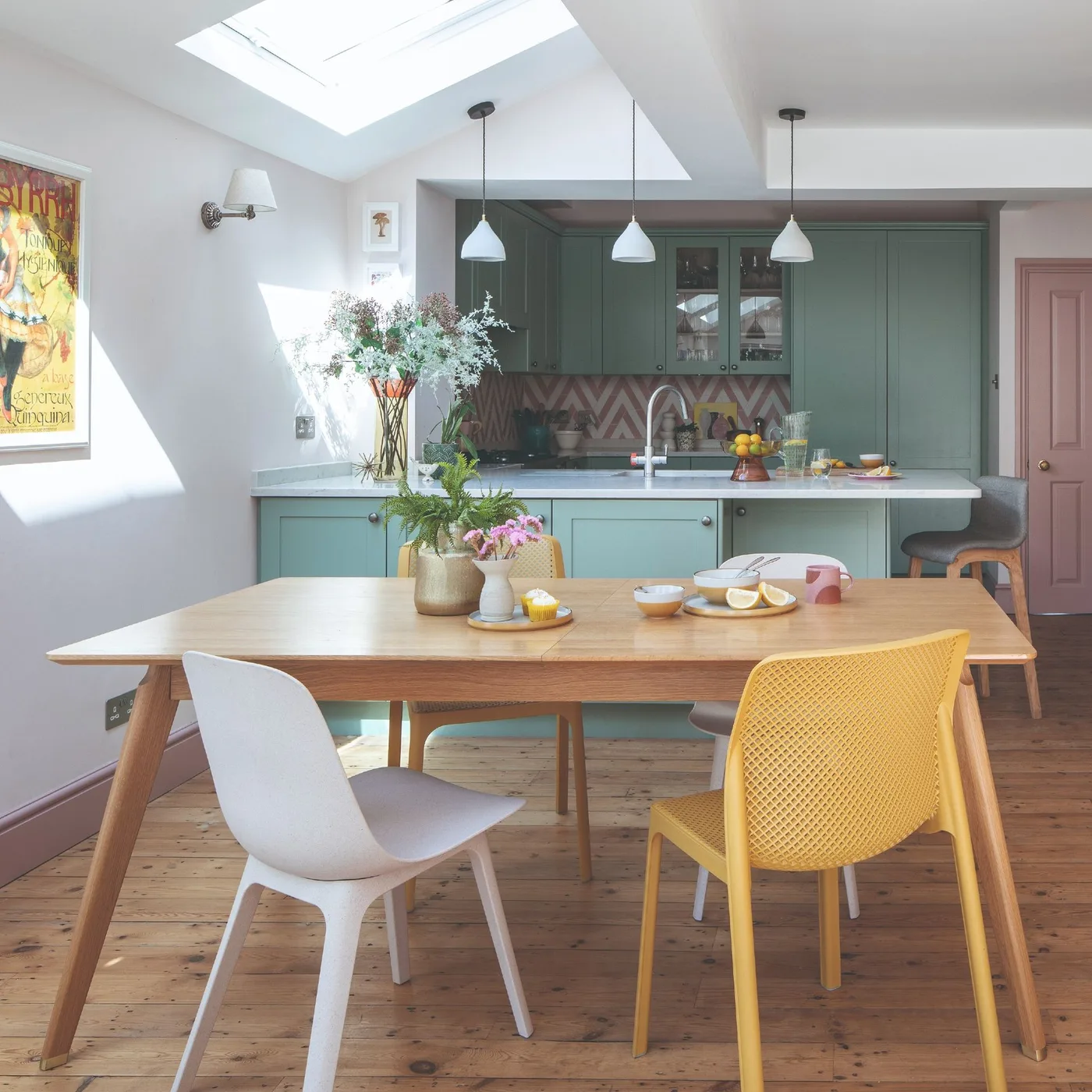 Open dining area with a wooden table, mixed yellow and white chairs, a green kitchen, pink door and pale pendant lights.