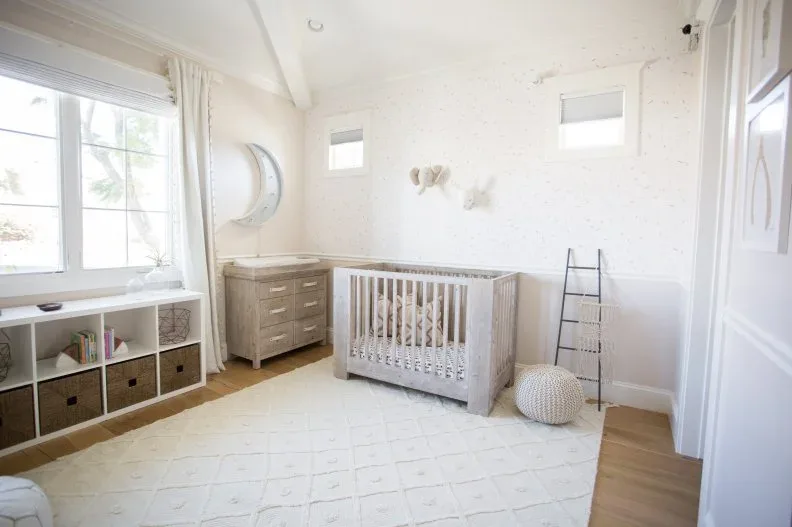 A pale wood nursery crib and dresser stand beside white walls and low basket storage under a window with a pale patterned rug on wood flooring