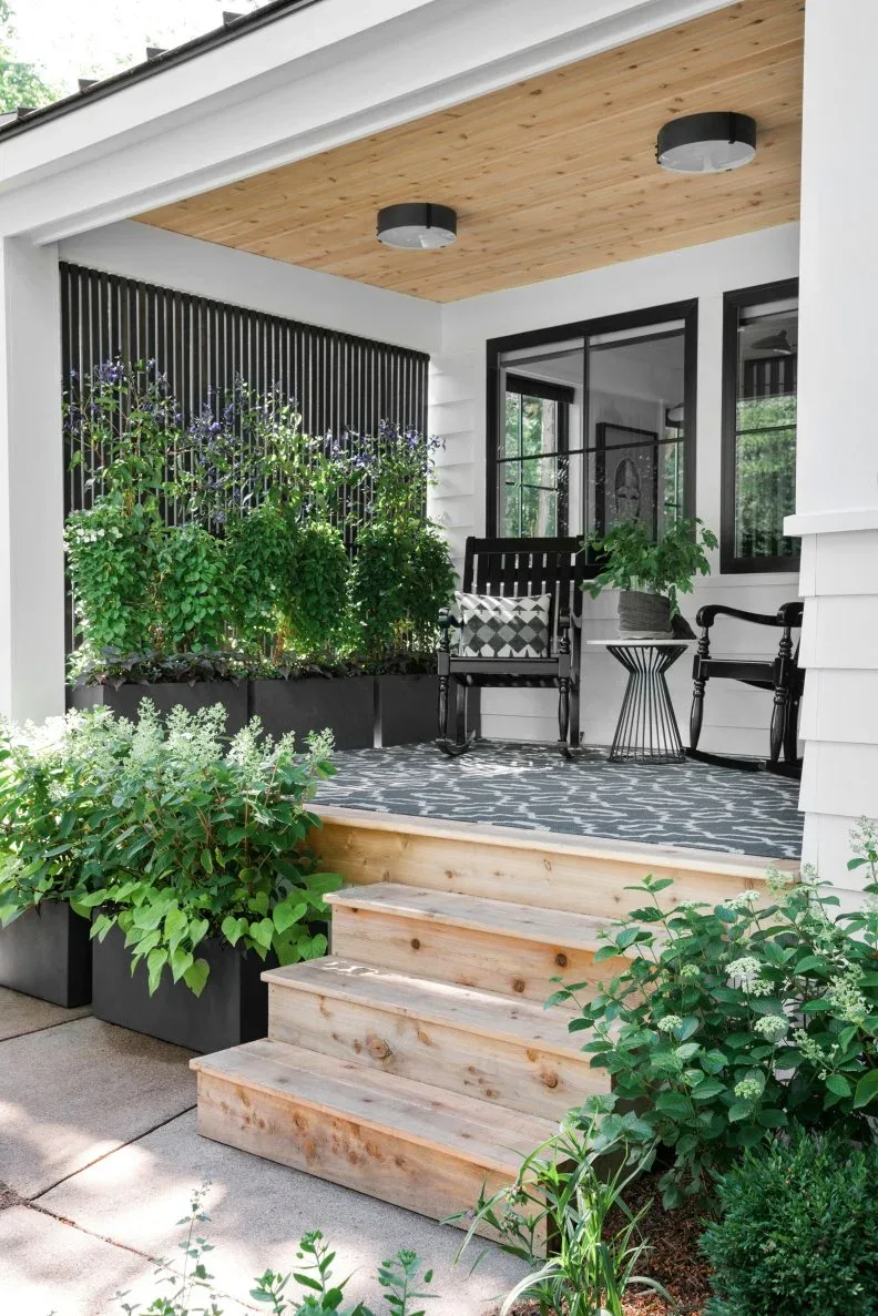 Small white porch with black rocking chairs, patterned rug, wood ceiling and black plant screen