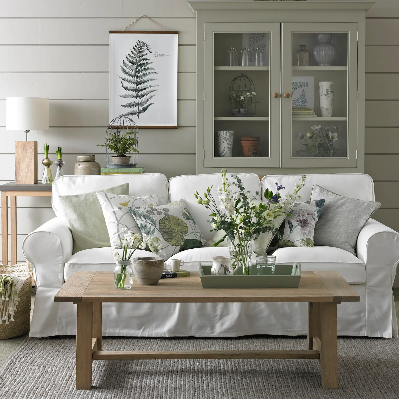 A white sofa with botanical cushions sits beside a wooden coffee table and pale green glass-fronted cabinet