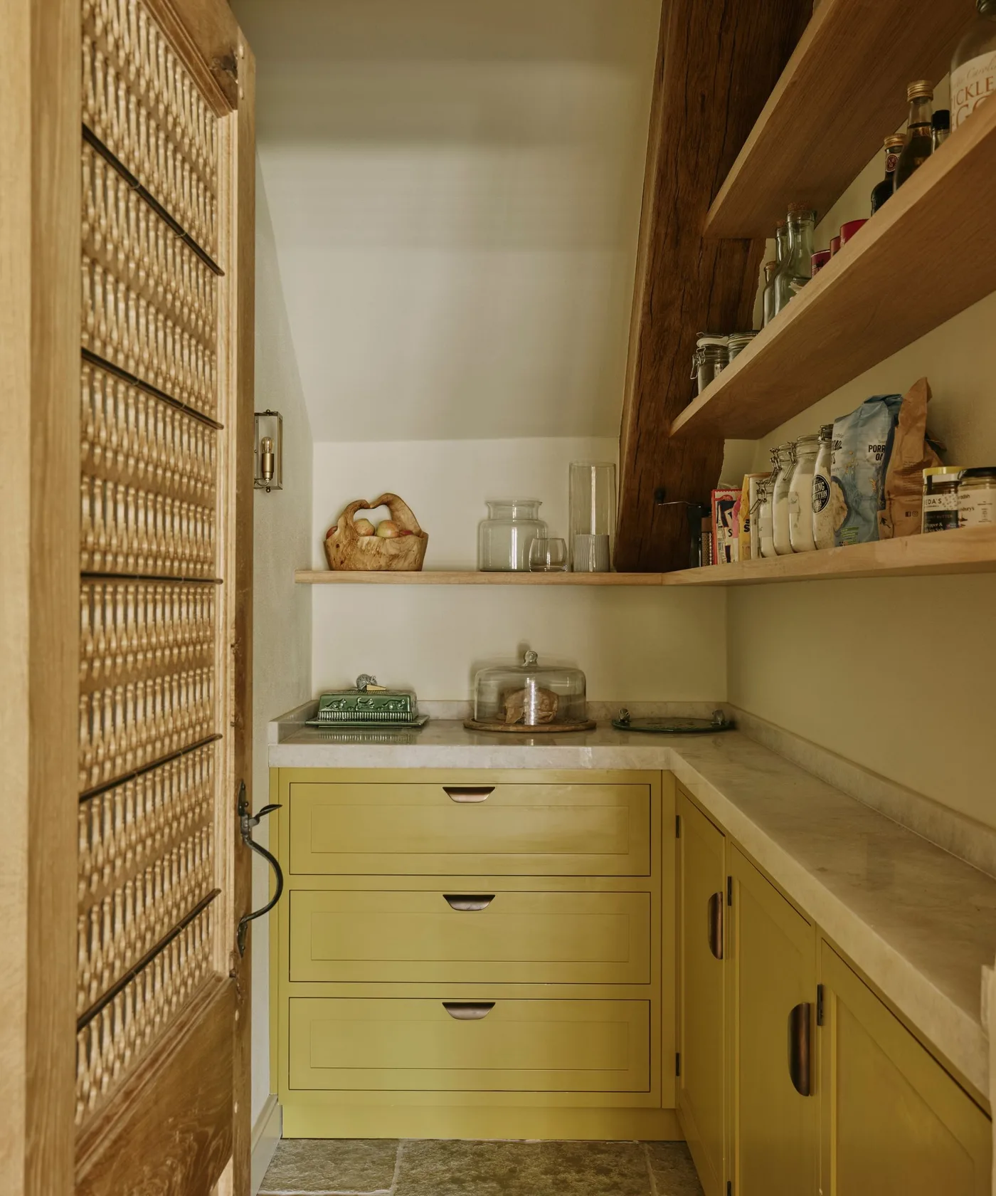 Understairs pantry with yellow drawers, wood shelves following sloped wall and textured open door