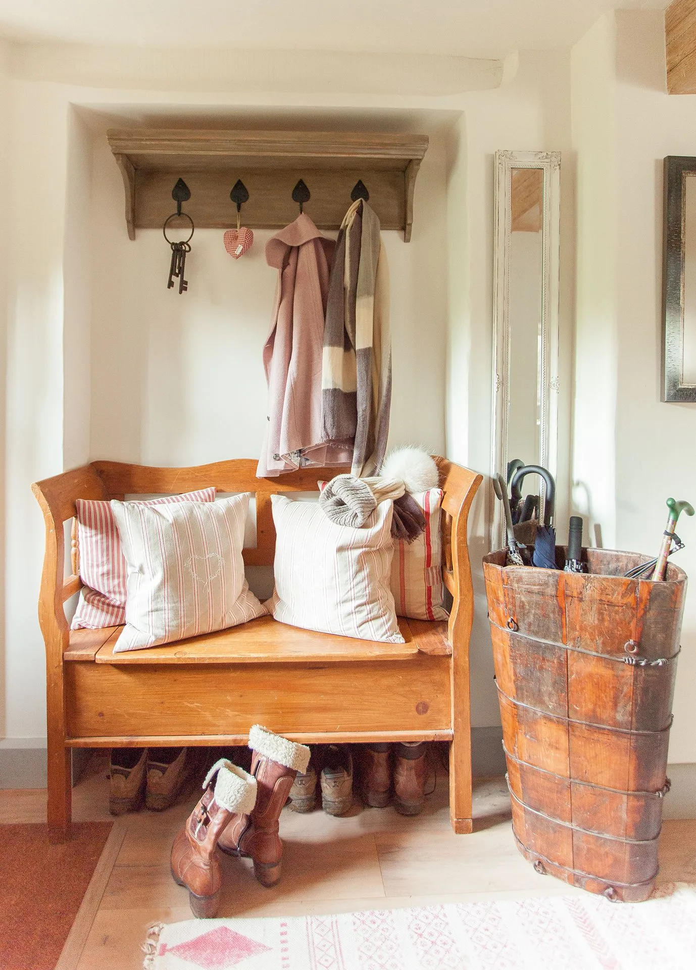 Country entrance with a wooden settle bench, pale cushions, coat hooks on a shelf rail, shoes below and a tall wooden umbrella holder.