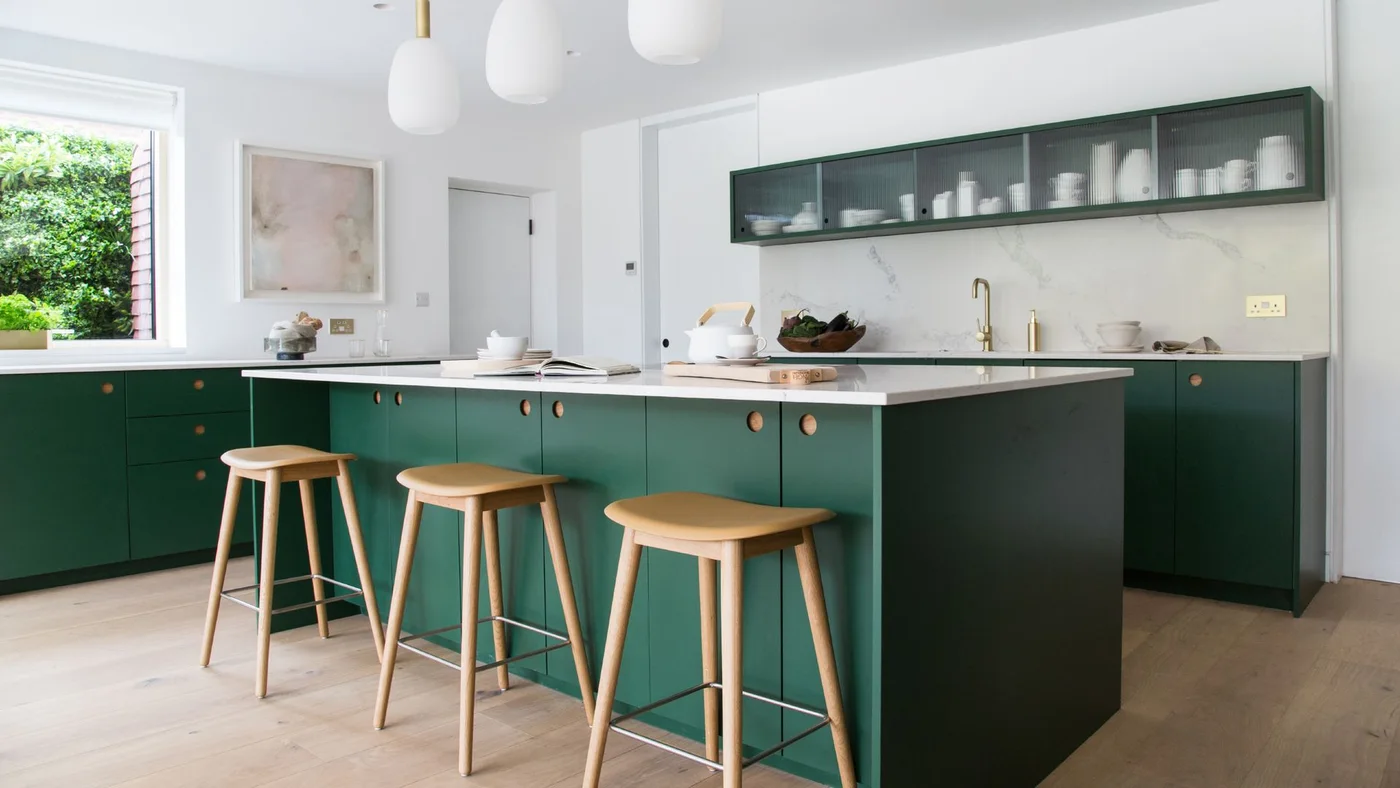 Green kitchen cabinets and an island sit below pale walls and a narrow glass-fronted wall cabinet