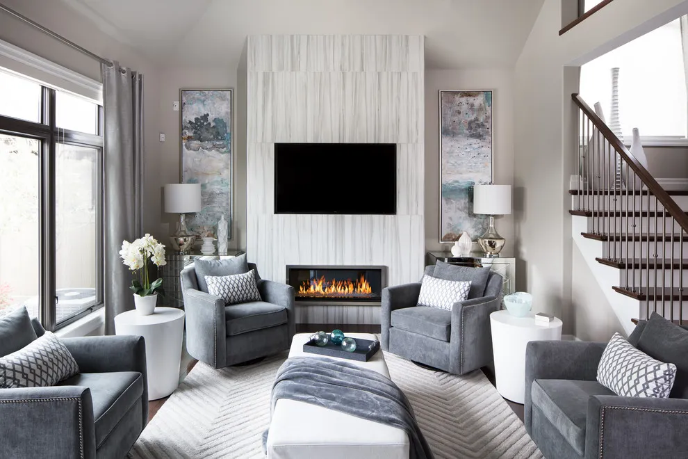 Four gray armchairs surround a pale ottoman in a tall room with a white fireplace