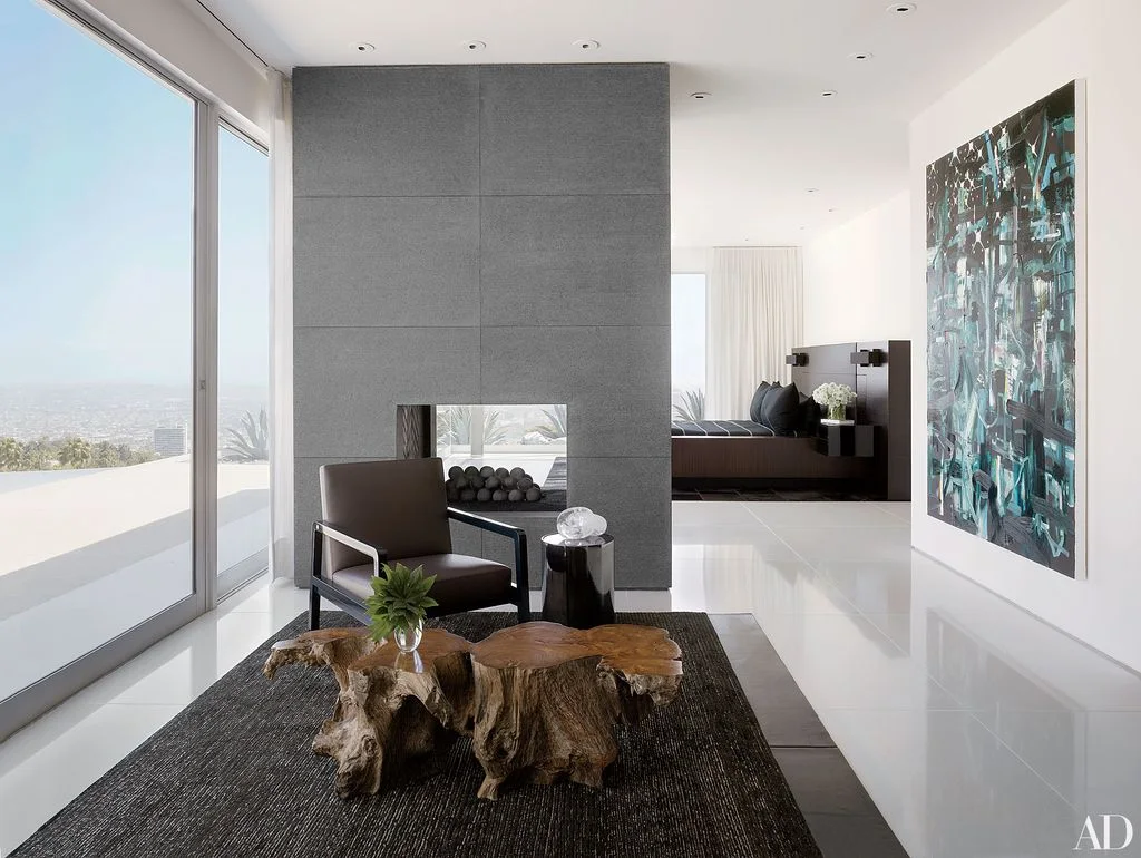A tall gray fireplace partition divides a sitting area from a bed beside large windows, a dark chair and sculptural wooden table