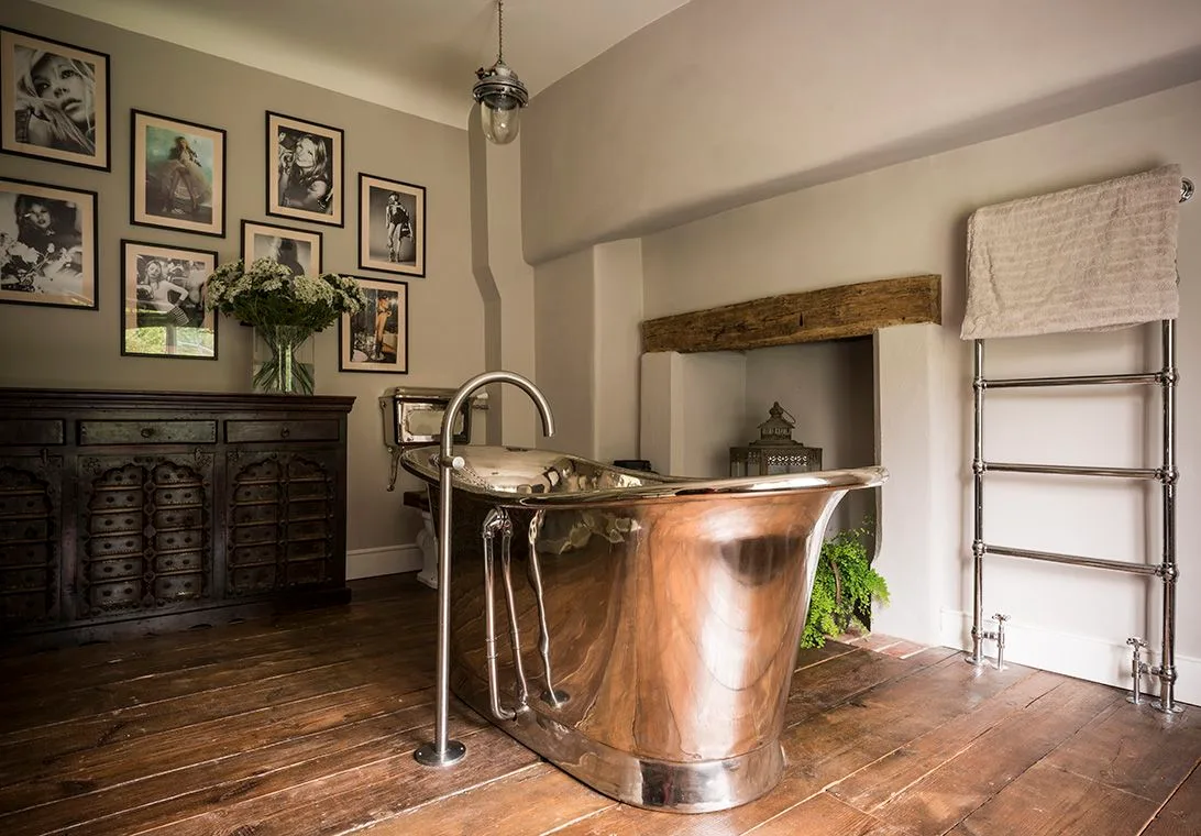 Bathroom with a warm metal freestanding bath, dark carved cabinet, framed pictures, wood floor and a small recessed fireplace opening.