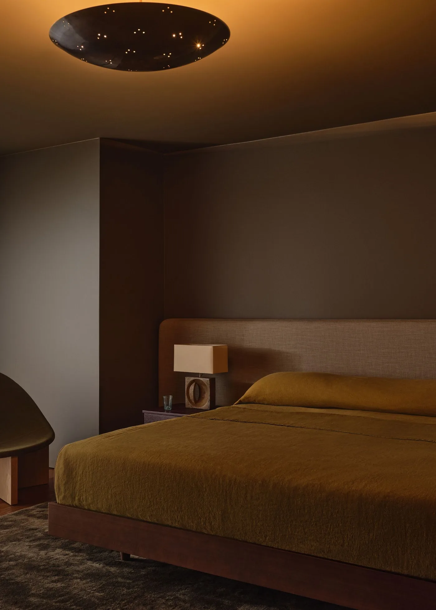 Deep brown bedroom with ochre bedding, a long dark headboard, a broad ceiling light and a cube-shaded bedside lamp