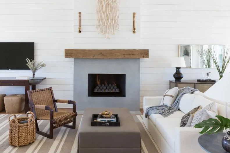 Plain gray fireplace with wood beam mantel against white horizontal boards, beside woven chair, basket and pale sofa