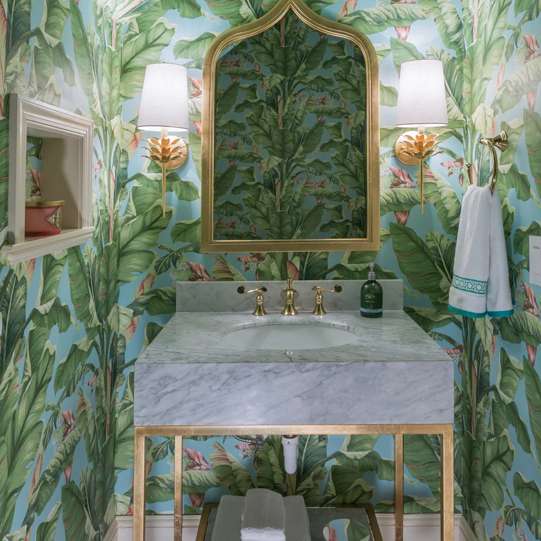 Tropical leaf wallpaper surrounds a curved gold-colored powder-room mirror, decorative shaded wall lights and a pale veined sink on an open stand