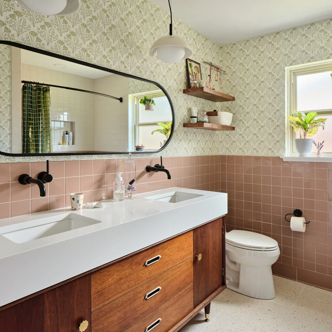Pink square tiles sit below pale botanical wallpaper beside a double sink, dark taps and wooden drawers
