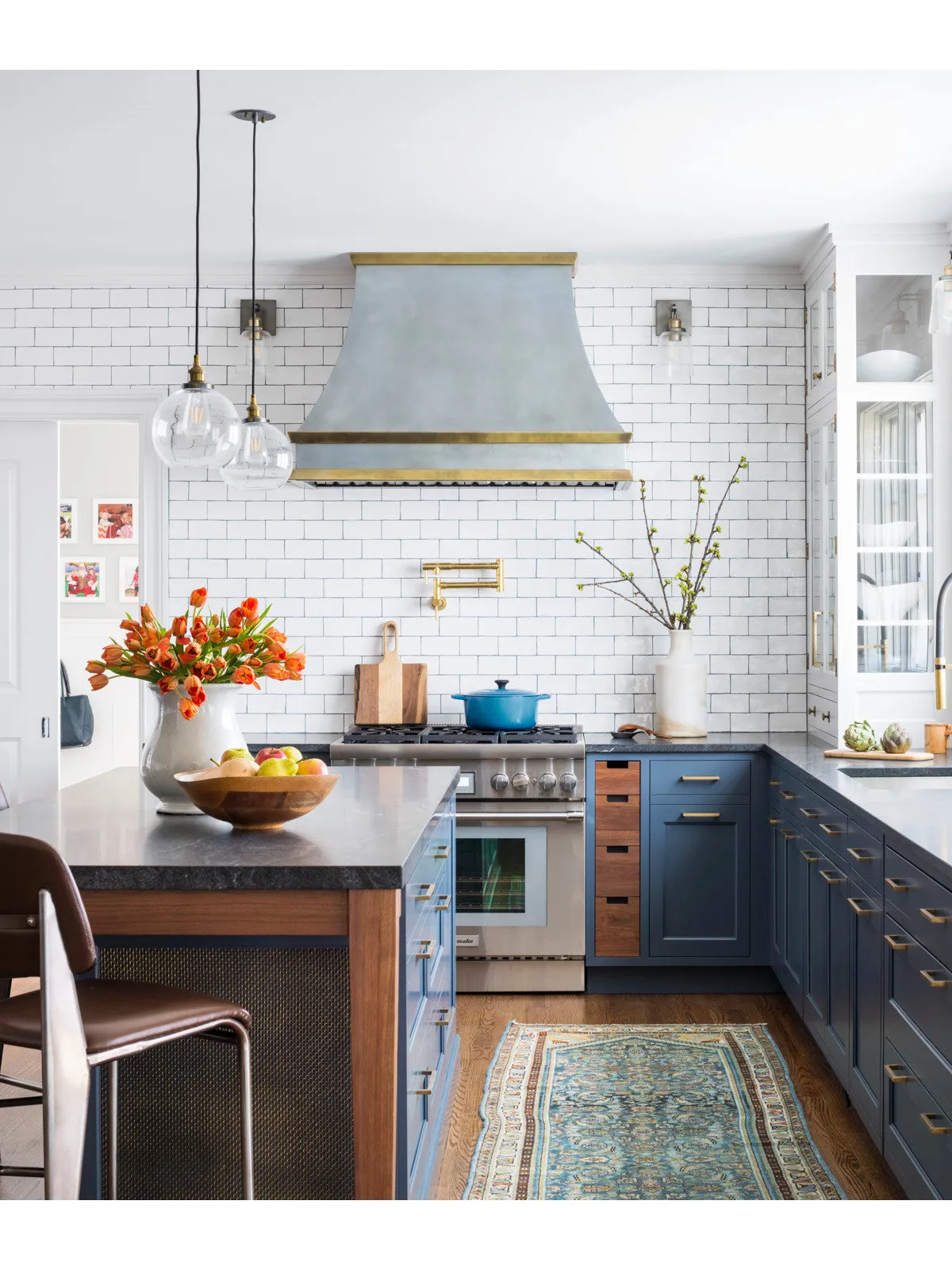 Navy base cupboards and drawers sit beneath a pale hood and white rectangular tiles beside a cooker, glass pendants and patterned runner