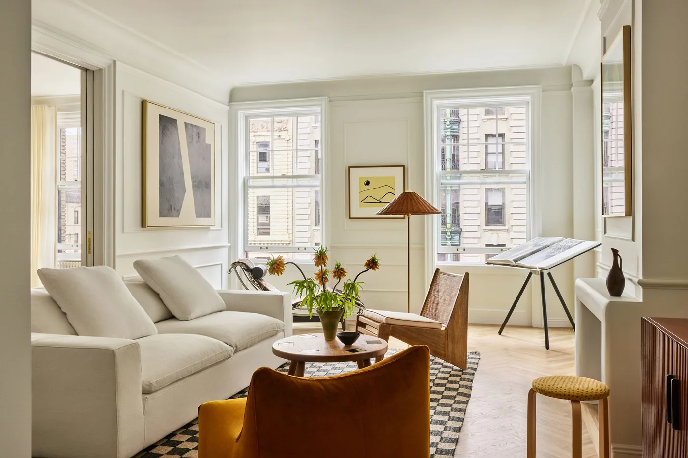 A pale sofa, brown accent chair and open-framed wood chair stand above a checked rug beside two tall windows and smaller framed artworks