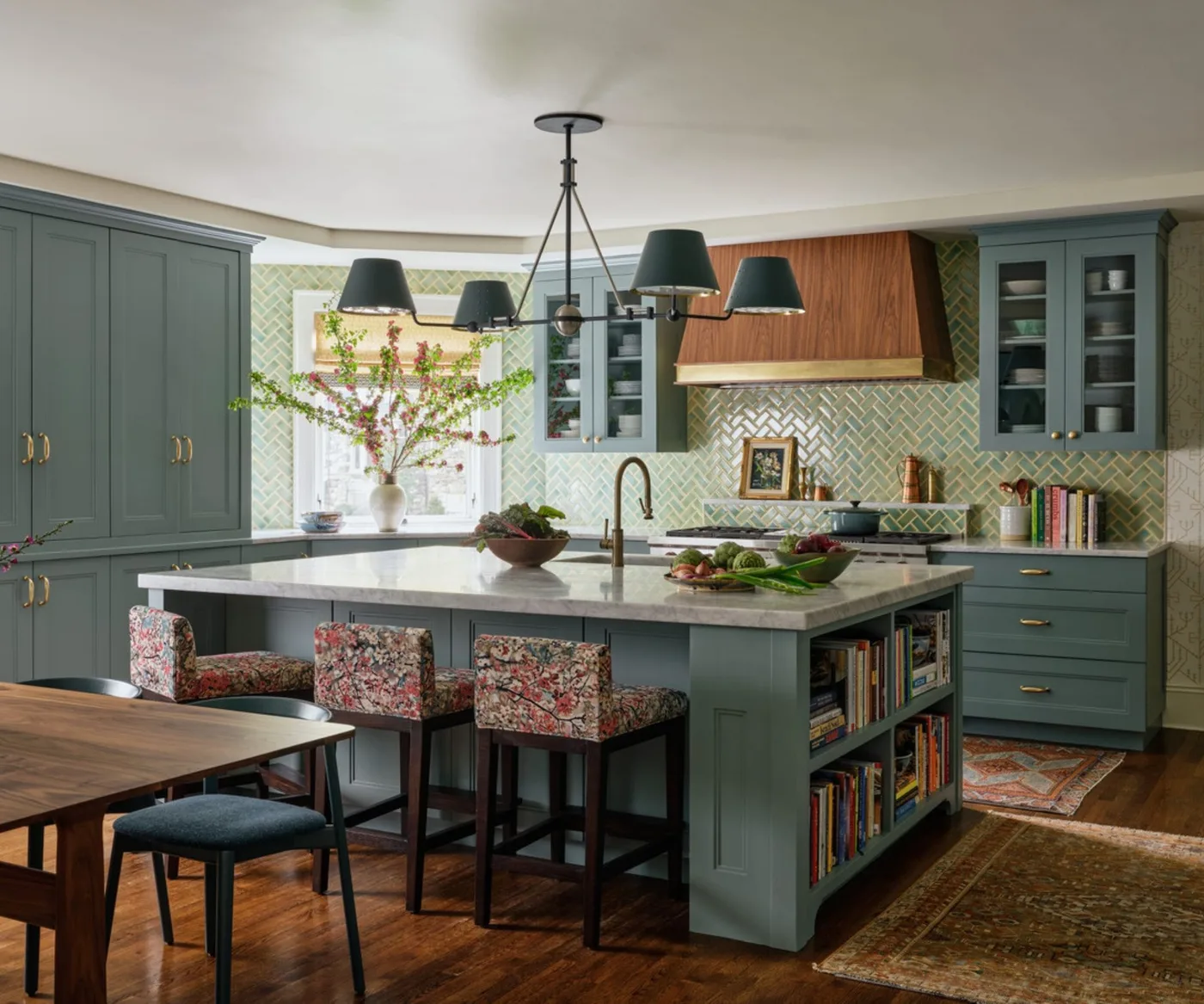 Blue-green kitchen cabinets with a pale green herringbone backsplash, wood-clad hood, patterned stools and a large island.