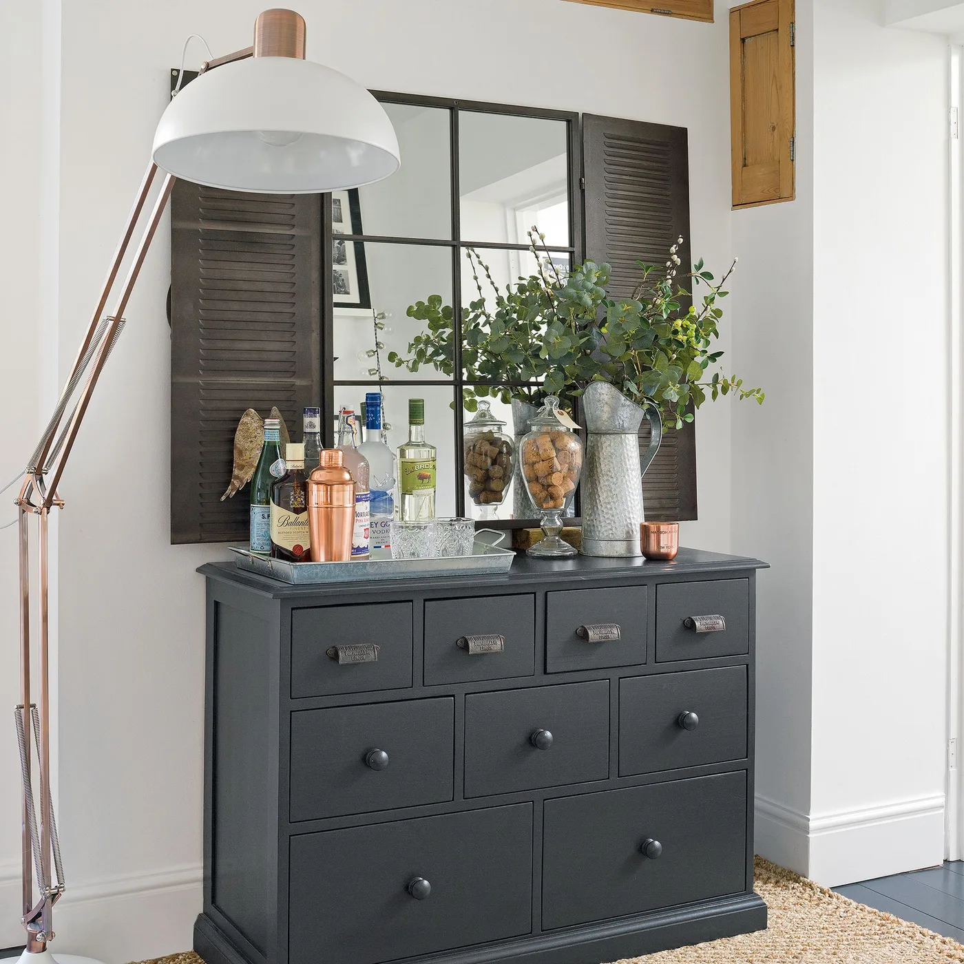 Dark shutter-style mirror above a gray drawer cabinet holding bottles beside a large pale floor lamp