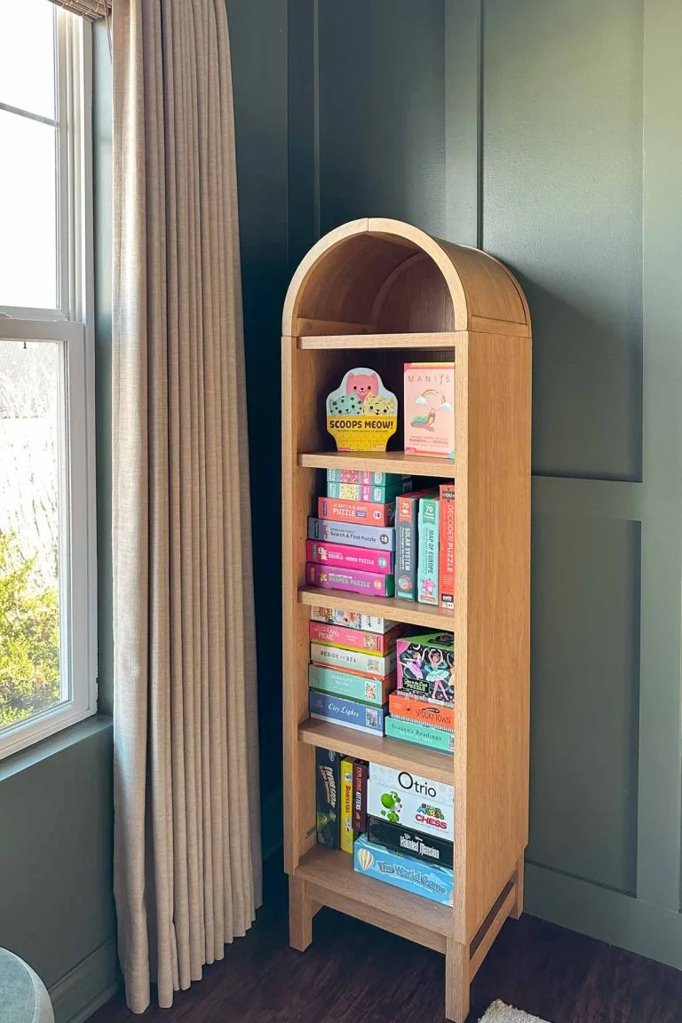 A warm wood-toned arched shelf holds books and board games beside a window, pale curtain and deep green paneled dining wall