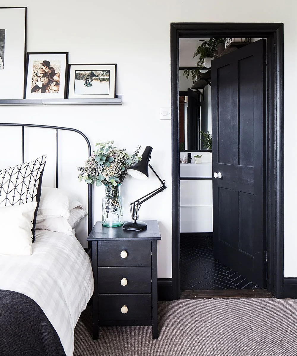 A dark bedside drawer chest and adjustable black lamp sit below framed pictures beside white bedding and a black door opening