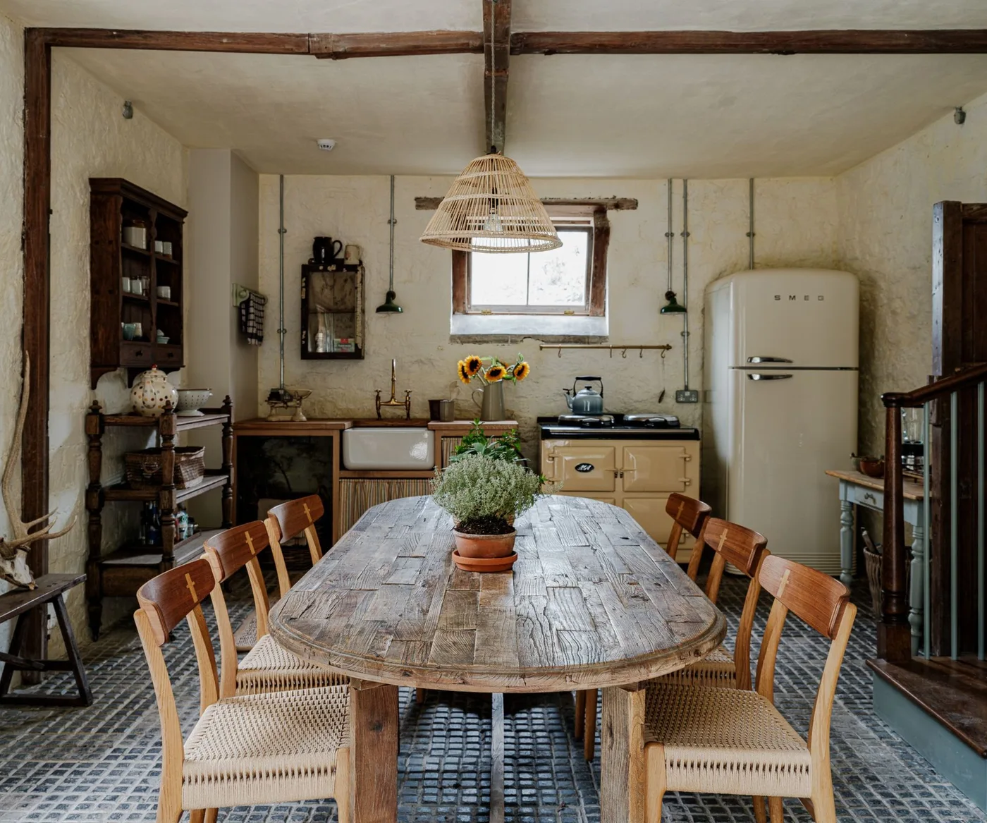 Cottage kitchen with a long rounded wooden table, woven-seat chairs, pale sink cabinet, cream cooker, tall pale refrigerator and exposed beams.