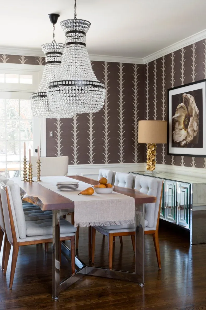 Two clear tiered chandeliers with dark bands above a rectangular wooden table and pale tufted chairs, beside dark floral wallpaper and mirrored cabinetry.
