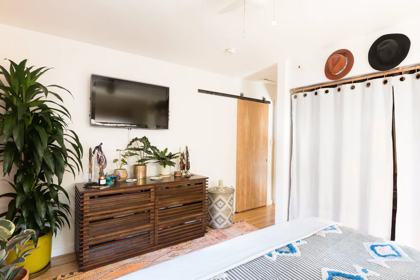 Bedroom with dark slatted wooden dresser, tall floor plant, smaller potted plants on top, white curtains and pale patterned bedding in foreground.