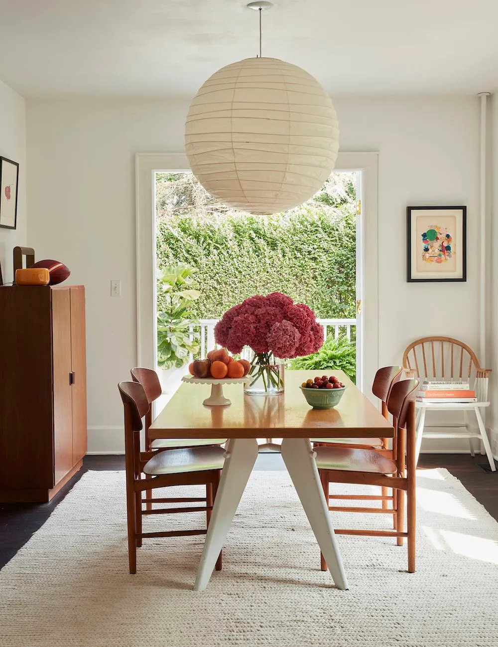 A small wooden table stands beneath a round pale pendant beside brown wooden chairs, a cabinet and a bright window