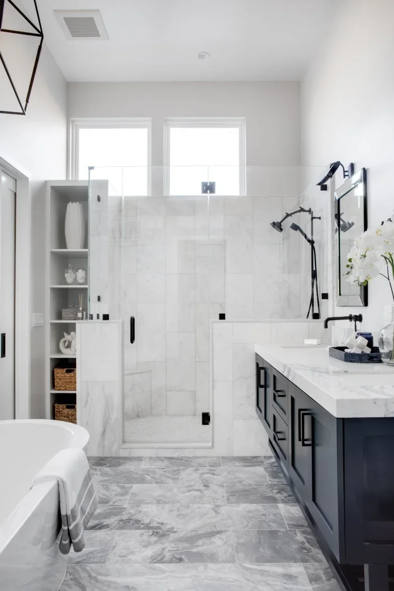 A pale bathroom has a dark vanity, white counter, glazed shower and open towel shelves beneath two small high windows