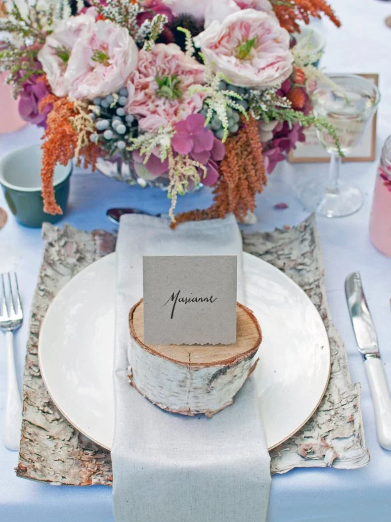A handwritten name card stands in a small wood slice above a pale plate and folded napkin with a bark-patterned mat below
