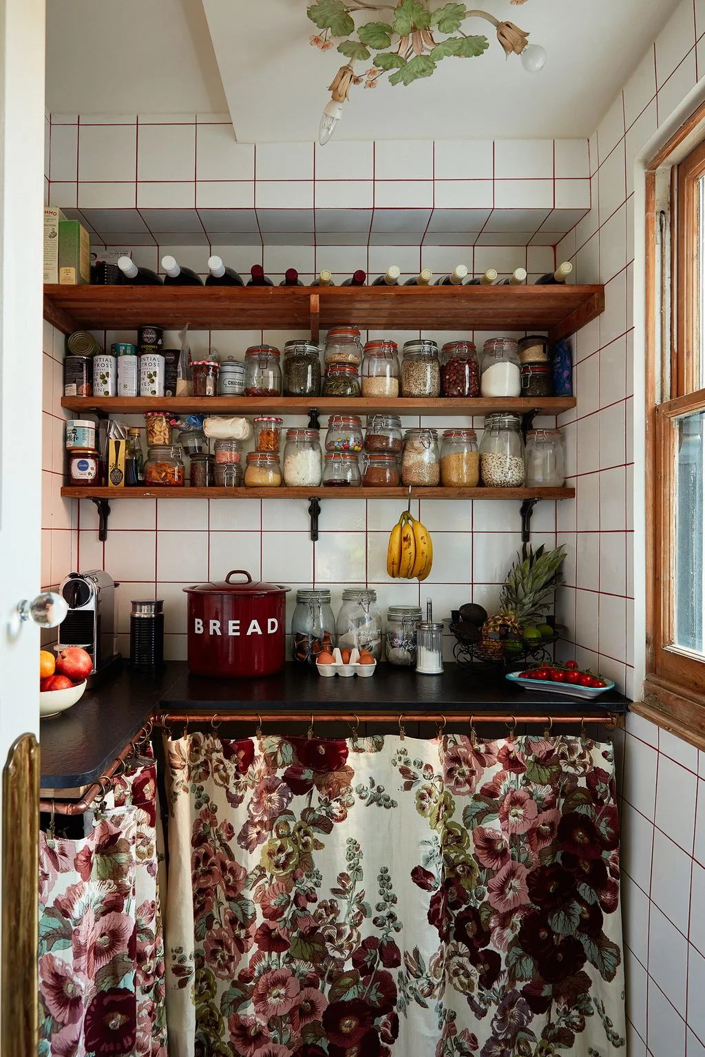 Open pantry shelves hold jars and bottles against white tiles with red lines above a floral cabinet curtain beneath a decorative branching light