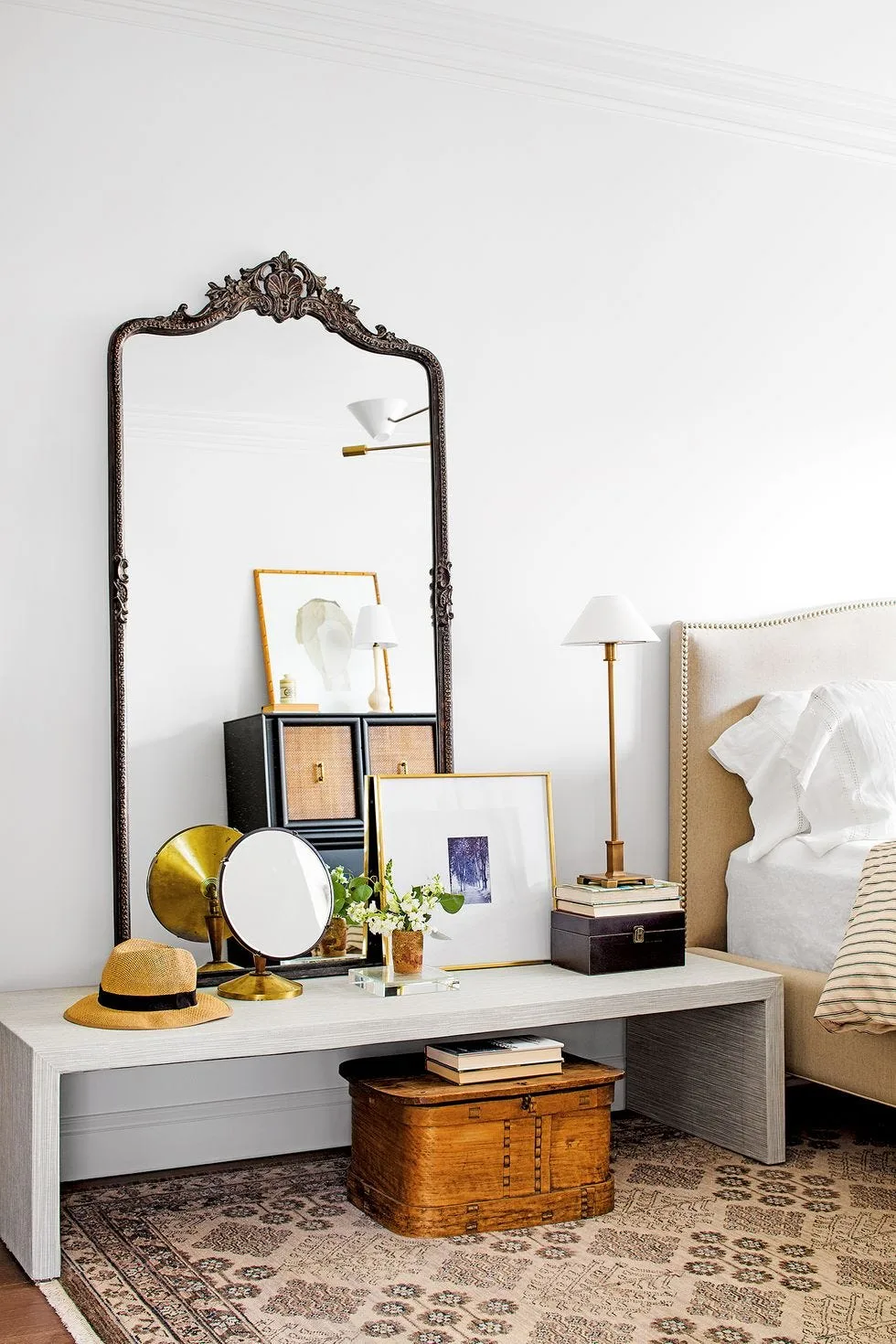 Bedroom with tall ornate dark-framed mirror, long pale console, smaller mirrors and framed pictures, wooden chest beneath and pale bed beside it.