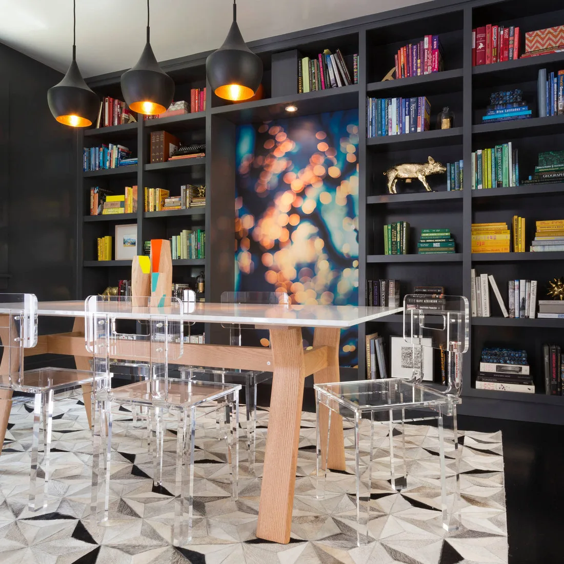 Pale table and clear chairs in front of black bookshelves with colorful books and three black pendants