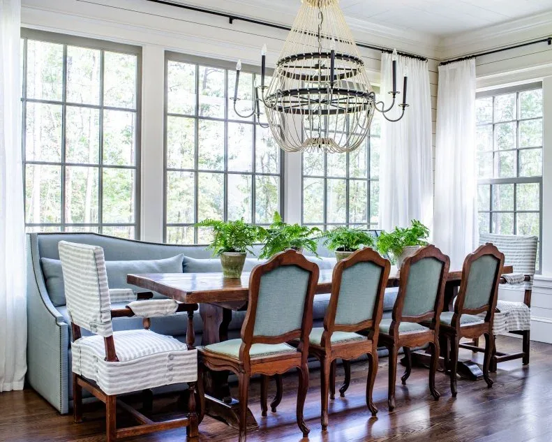 Several green potted plants along a long wood dining table with blue chairs and a window bench